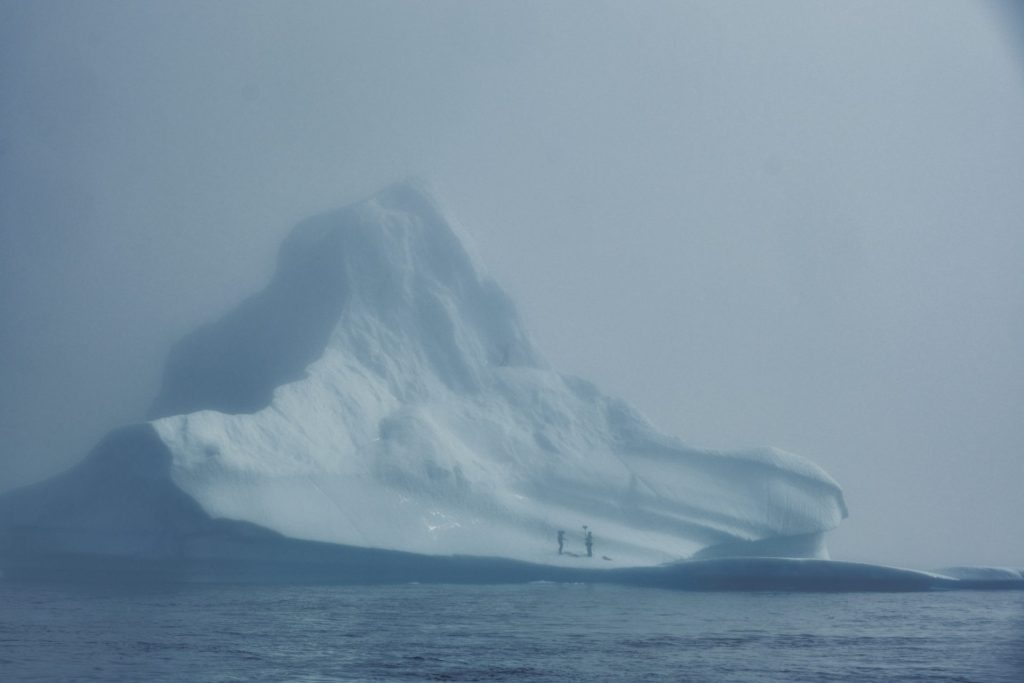 Image of iceberg looming in the mist, and two silhouetted figures standing upon it.