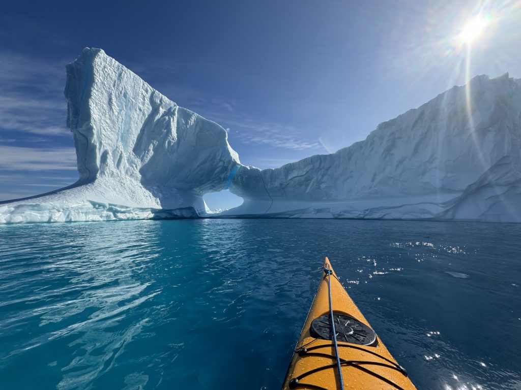 A kayak paddles toward a dramatic iceberg, pierced with a hole through its center.